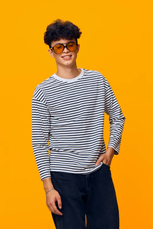 young man wearing striped shirt and sunglasses, smiling with hand in pocket against bright orange background, casual style and joyful mood