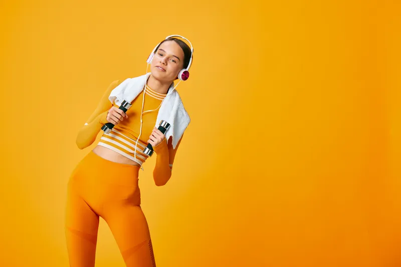 Fitness woman in bright orange sportwear stands with dumbbells, towel and headphones, posing confidently against a vivid orange backdrop for energetic workout lifestyle.