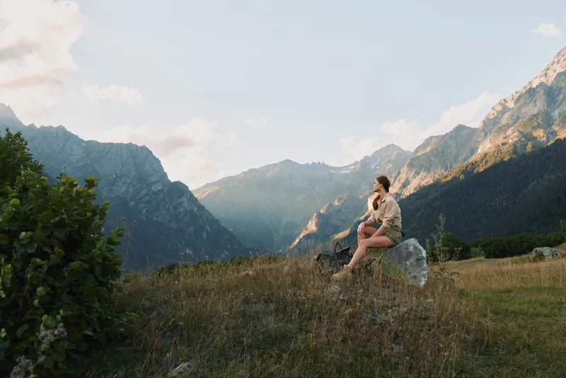 Calm woman sits on a rocky outcrop in a meadow among mountains, embracing outdoor life. Soft light and expansive scenery convey wellness, relaxation, and natural landscape in a simple portrait.