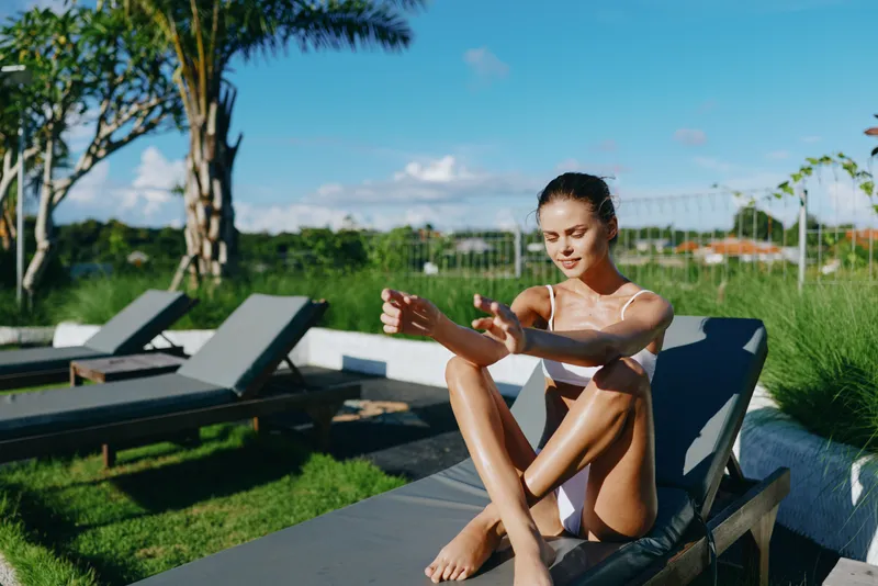 Relaxed woman in a swimsuit sits on a poolside lounge applying sunscreen. Bright outdoor setting with palm trees, green lawn, and blue water. Lifestyle scene focusing on skincare, wellness, self