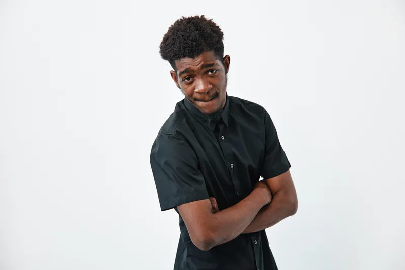 Young African American man with dark skin and curly hair wearing black shirt standing with crossed arms on white background looking at camera with confident expression.