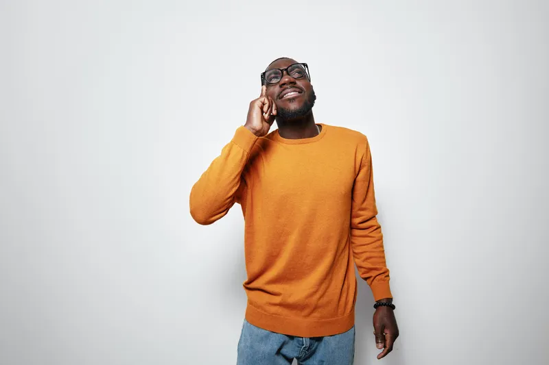 African American man with dark skin wearing orange sweater and glasses talking on smartphone smiling isolated on white background. Casual style and positive emotion.
