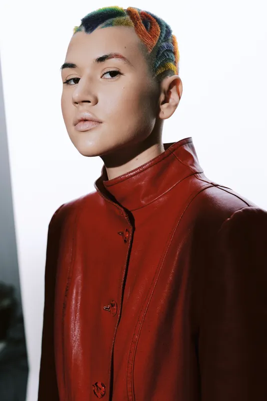Young woman with colorful short hair wearing a red leather jacket, looking confidently with neutral expression, contemporary fashion portrait in studio lighting.