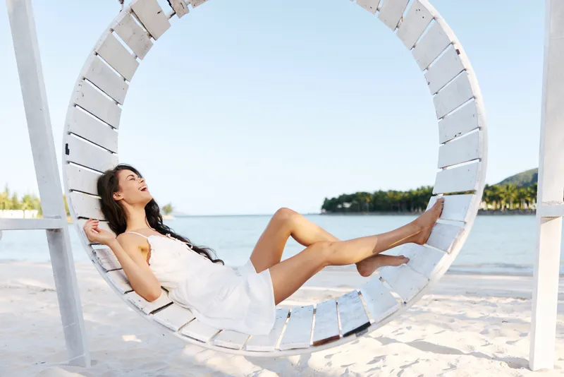 A woman rests on a circular wooden swing on a white sandy beach, gazing toward the sea, sunlit calm, capturing a serene moment of coastal leisure.