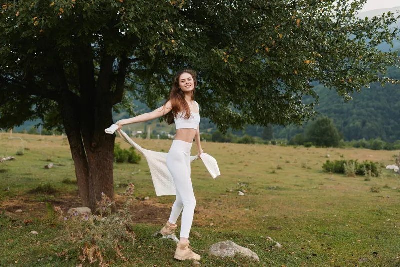 Young woman in white sportswear enjoys nature near tree in open field with mountains background, smiling and holding jacket, healthy lifestyle concept outdoors.