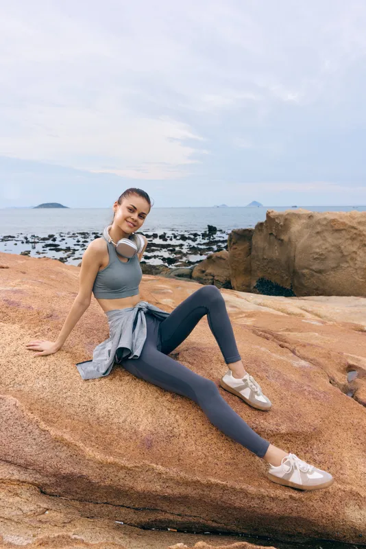 Happy fitness woman sitting on a rocky beach, wearing activewear, enjoying a healthy lifestyle, with headphones and a calm ocean backdrop