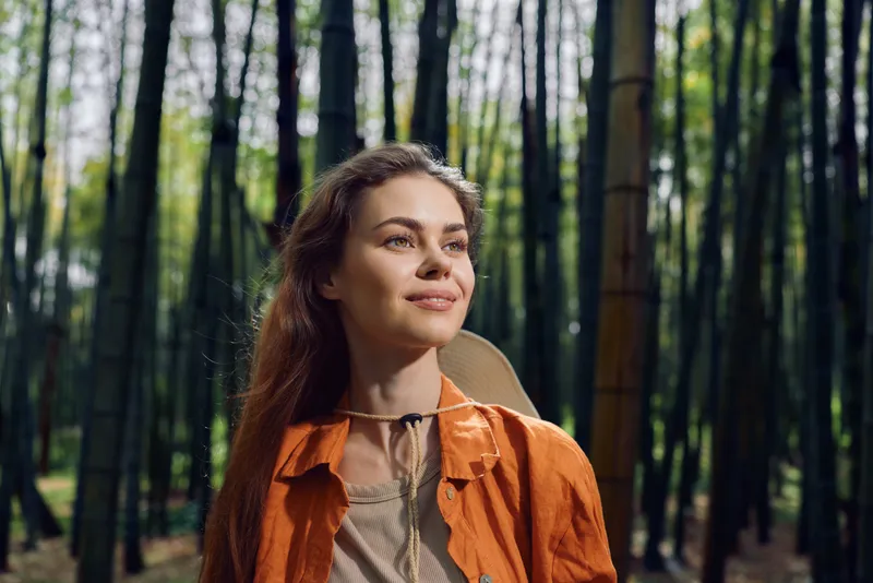 Woman portrait smile in a bamboo forest, nature outdoors scene with soft sunlight on her face, relaxed expression and casual orange jacket conveying peaceful hiking and leisure.