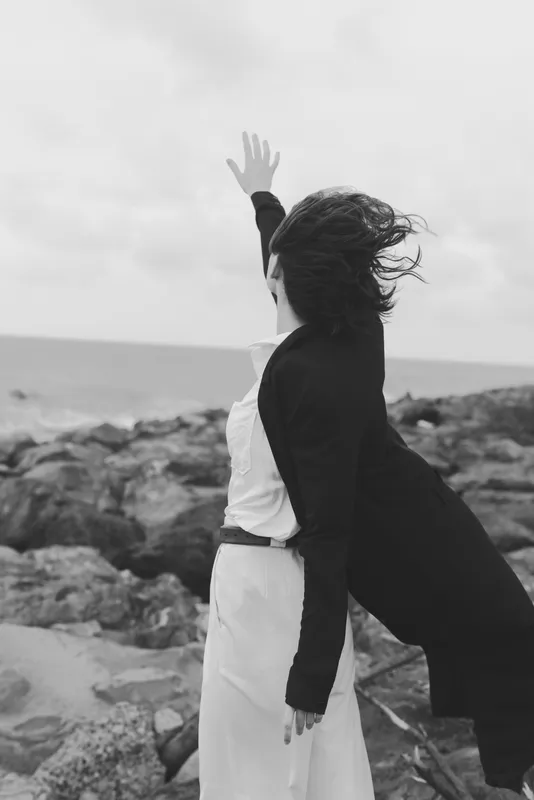 Silhouettes on a windy beach, two figures in contrasting outfits raise arms against a rugged seascape, capturing motion, freedom and connection in a monochrome scene