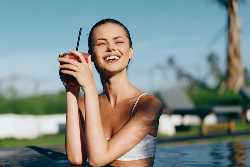 Smiling woman by an outdoor pool drinks a cold beverage under bright sunshine. A relaxed summer lifestyle scene focused on hydration, leisure, and warmth beside clear blue water.