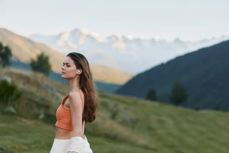A cheerful woman stands in a bright outdoor landscape with rolling hills and distant mountains. She wears an orange crop top, enjoying the fresh air and peaceful scenery.