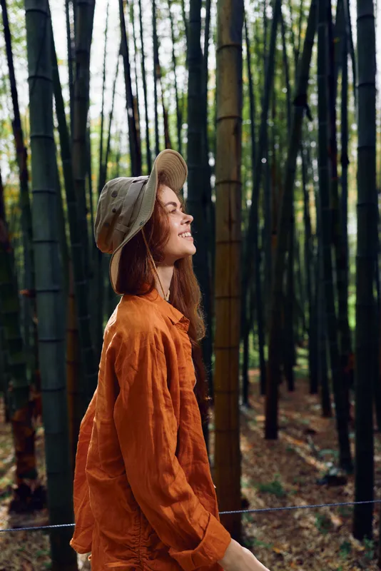 Woman smile hat bamboo forest portrait nature outdoor scene with orange jacket, serene travel moment walking among tall green stalks and dappled sunlight in peaceful grove.