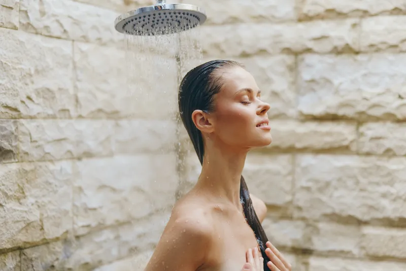 Young woman enjoying outdoor shower with eyes closed, wet hair, and serene expression against beige stone wall, natural light creating peaceful atmosphere.