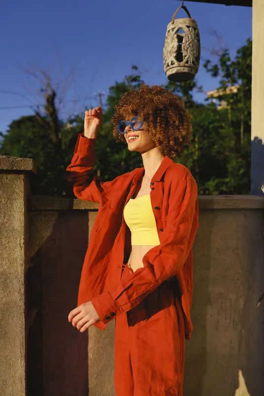 Smiling woman with curly hair wearing a red jacket and yellow top outdoors, posing on a terrace. Concept of lifestyle, fashion, and confidence in warm sunlight