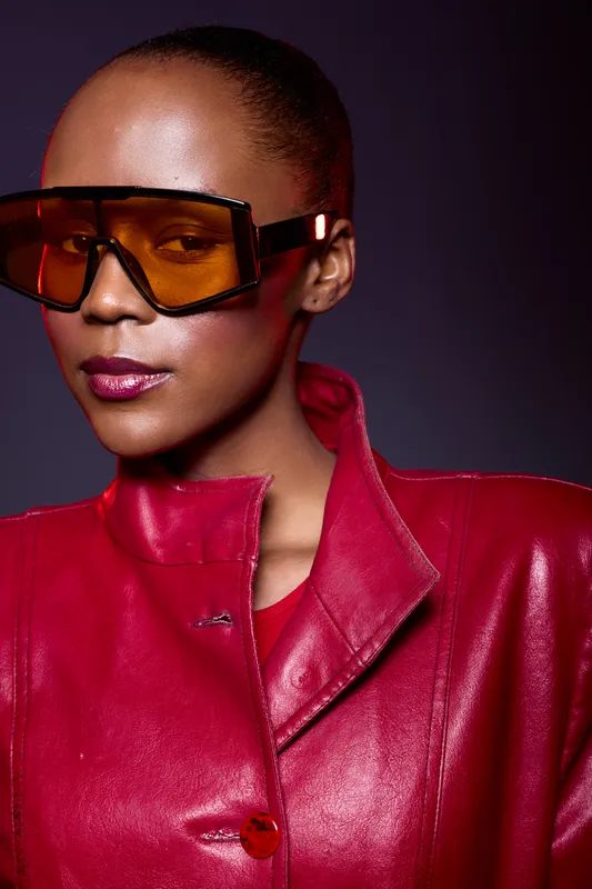 Elegant African American woman with dark skin wearing oversized orange sunglasses and a shiny red leather jacket. Studio portrait with dramatic lighting and confident expression.