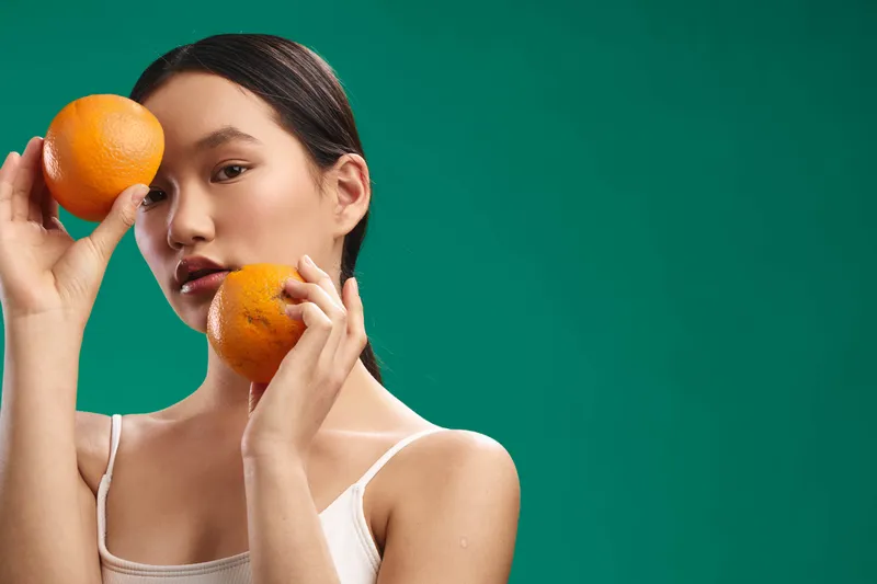 asian woman is photographed in a studio setting, holding bright oranges near her face against a vivid green backdrop. she gazes softly, exuding freshness, confidence, and a playful healthy lifestyle.