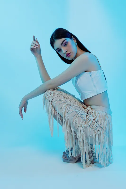 Young woman in white crop top and fringed pants poses against blue background. Fashion concept highlighting beauty, style, and modern clothing trends in studio lighting.