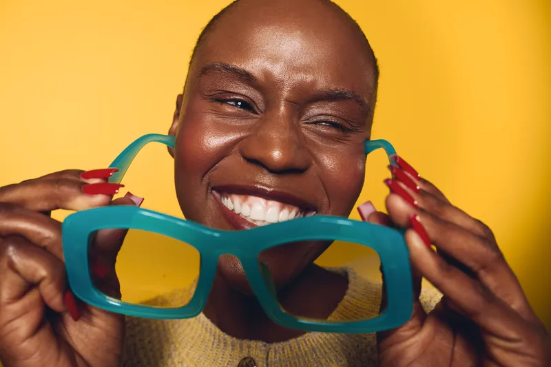 Close-up of smiling dark skinned African woman holding oversized blue glasses in front of yellow background. Concept of joy, fashion accessories, beauty and positive emotions.