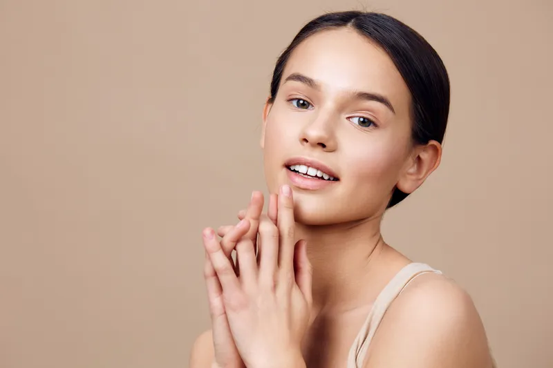 Young female model with a natural smile, showcasing healthy skin and minimal makeup against a soft beige background, representing beauty and confidence