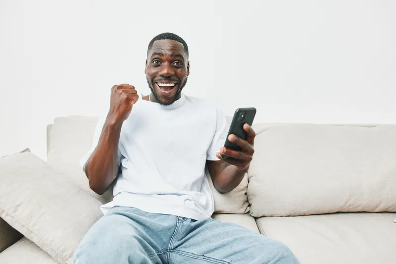Happy African man celebrating a victory while holding his smartphone, wearing casual clothes, on a cozy couch with a light background