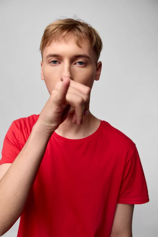 Shushing gesture with hand raised to lips by a man in a red shirt, studio setting, focused expression, conveying silence, secrecy, or quiet instruction for communication and mood in media