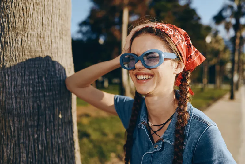 Smiling woman with blue sunglasses and red headscarf enjoys sunny day outdoors in casual denim outfit, expressive joyful mood, natural park background.