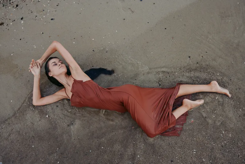 A graceful woman in a red dress lies on sand, posing softly with arms raised and distant gaze, creating a calm, dramatic still life on a beachlike surface.
