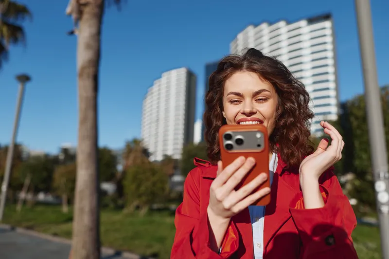 A happy young woman in a red jacket takes a selfie outdoors in a sunny urban park with tall buildings and palm trees in the background, enjoying a bright day.