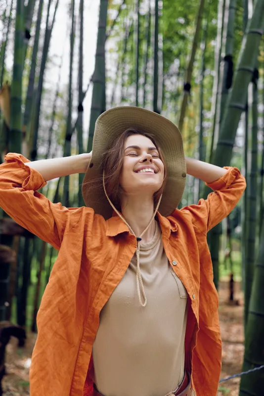 Woman with a relaxed smile wearing a wide brim hat in a dense bamboo forest, enjoying nature outdoors. Portrait of relaxation and sunlight on greenery for travel and lifestyle.