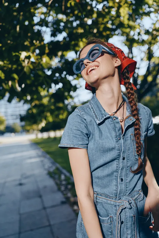 smiling woman in denim dress wearing sunglasses and red headscarf outdoors in sunny park with green foliage background enjoying warm weather