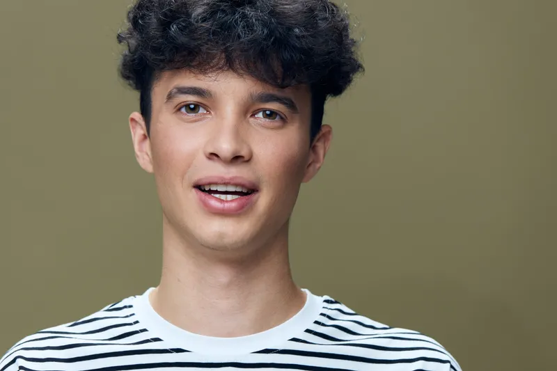 Young man with curly hair wearing striped shirt smiles softly with open mouth against neutral olive background, expressing friendly and approachable mood in studio setting.