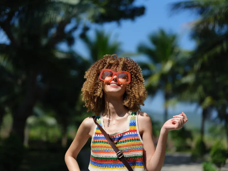 Smiling woman with curly hair wearing colorful sunglasses and a striped crochet top outdoors in sunny tropical environment.