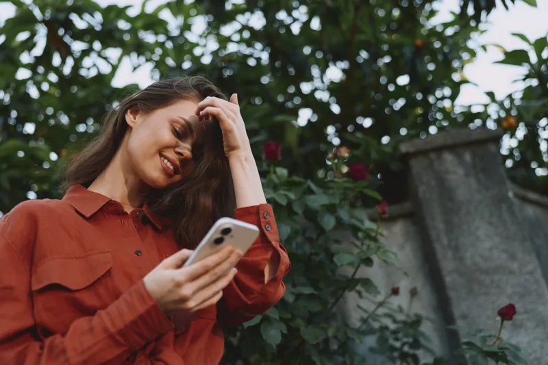 Smiling woman in a red jacket checks her smartphone outdoors, surrounded by greenery. Casual lifestyle moment highlighting connection, nature, and daily routines in natural light