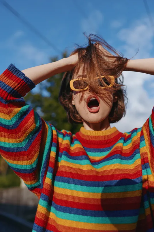 Woman in rainbow sweater and yellow sunglasses with playful expression, young adult outdoors with hands up, casual outfit and messy hair, vibrant fashion lifestyle portrait.