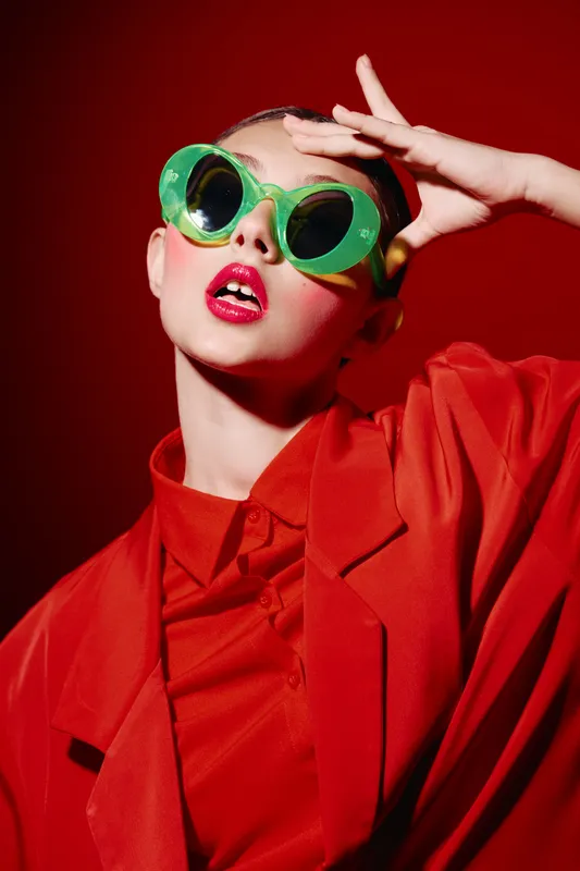 Fashion model wearing vivid red jacket and green oversized sunglasses poses confidently against a red background to emphasize beauty, creativity, and avant-garde style themes.