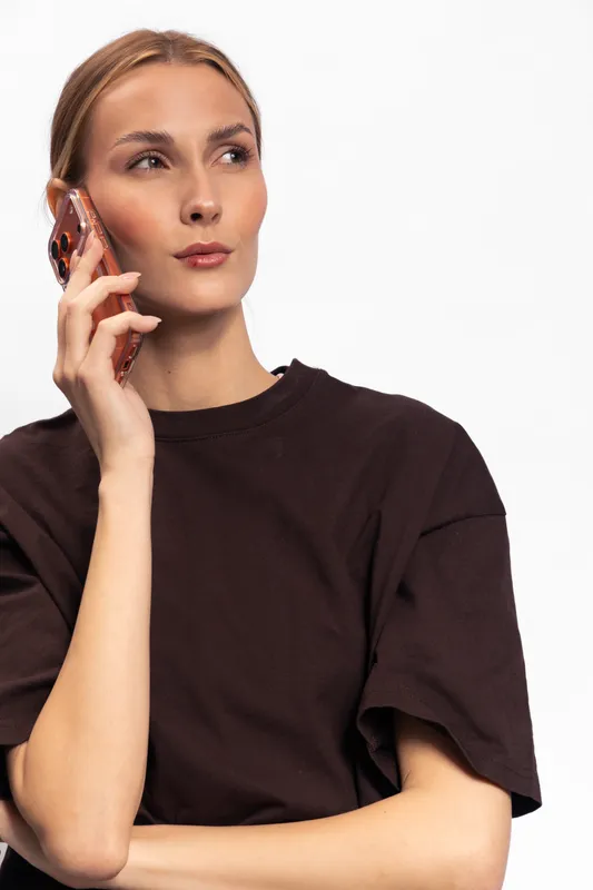 Young woman in brown t shirt holding smartphone near ear looks away thoughtfully on white background. Concept of communication, technology, and casual lifestyle.