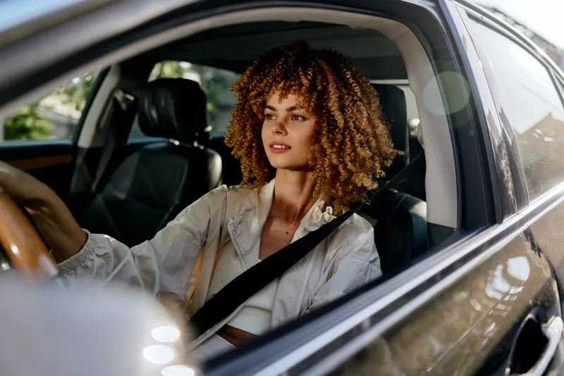 young woman driving car confidently with seatbelt fastened, natural curly hair, casual outfit, bright daylight, modern vehicle interior, safety conscious