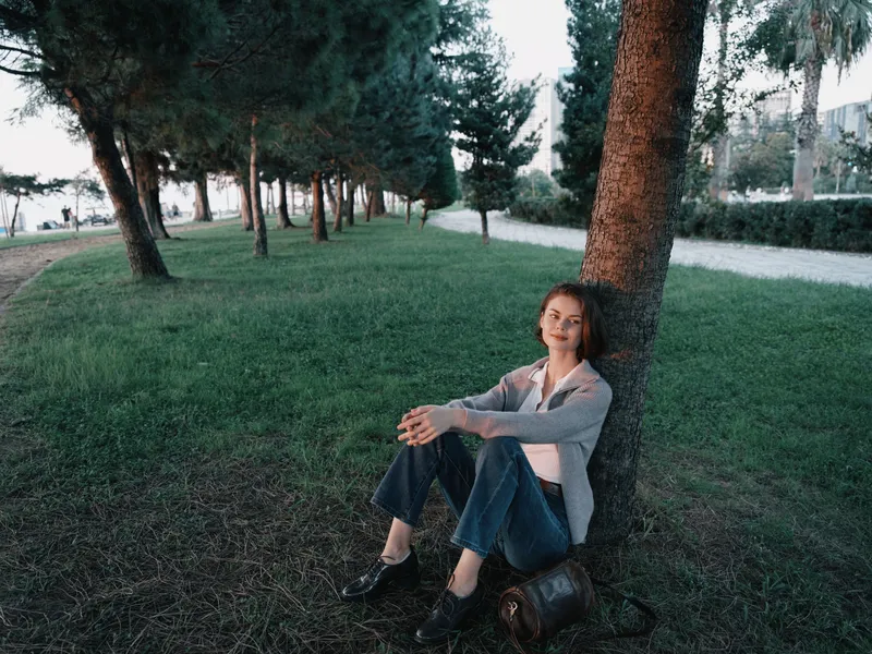 A calm professional woman sits against a tree in a green park, wearing smart casual attire. She relaxes outdoors, appearing thoughtful, composed, and focused in a peaceful moment.