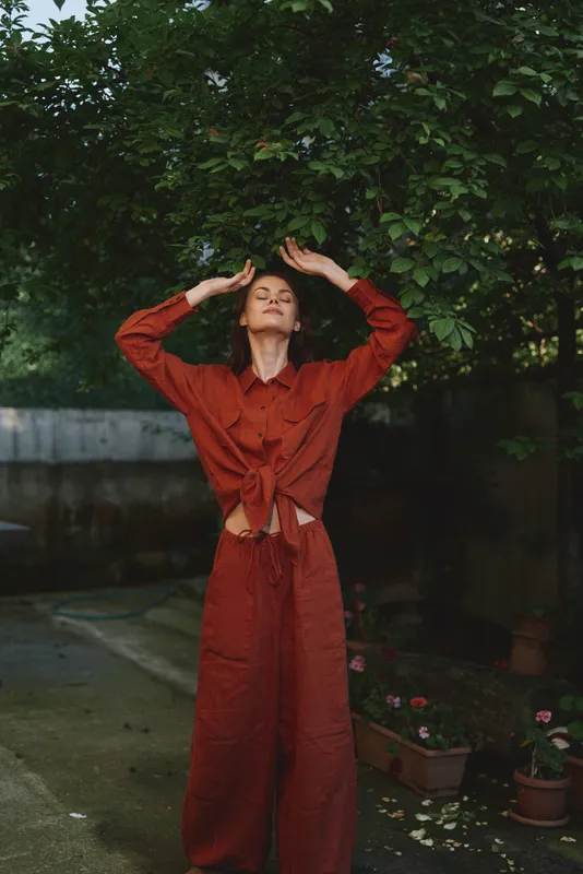 Sunlit garden scene shows a young woman in a red jumpsuit stretching with arms raised, conveying confident relaxation and a carefree mood
