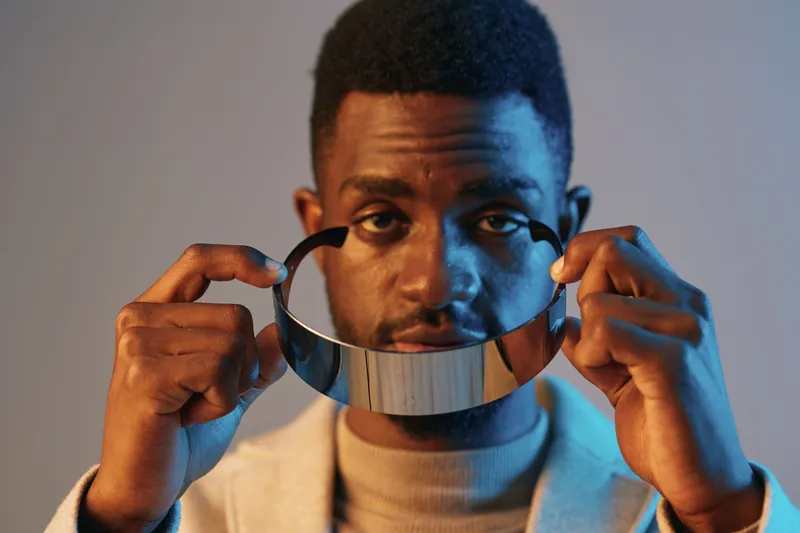 A focused Black man holding a reflective silver collar against a soft blue background, expressing curiosity and contemplation, blending modern fashion with artistic style
