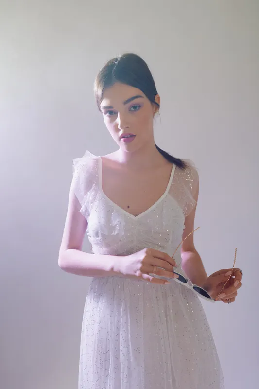Young woman in a white elegant dress poses softly against a pale background. Her makeup is natural, and she holds sunglasses in one hand. Concept of beauty, fashion, and elegance.