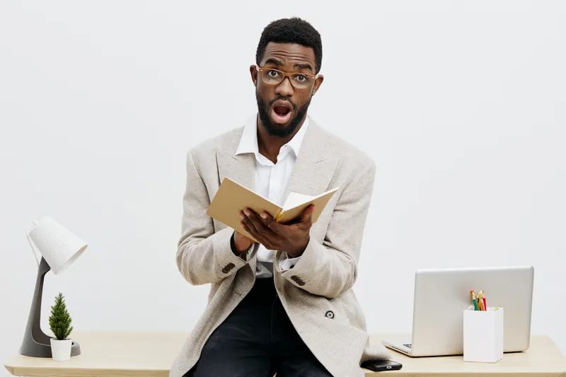 Confused young African American man in beige blazer and white shirt holding a notebook with surprised facial expression. Office or home workspace with laptop, desk lamp, and plant in background. Conce