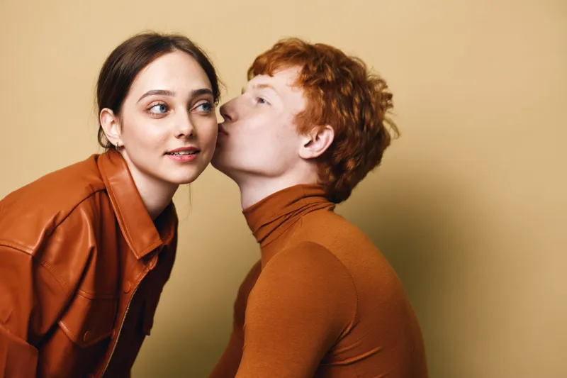 Man and woman in brown clothing, with man kissing woman on cheek against beige background. Couple showing affection, red hair and casual style with intimate moment.