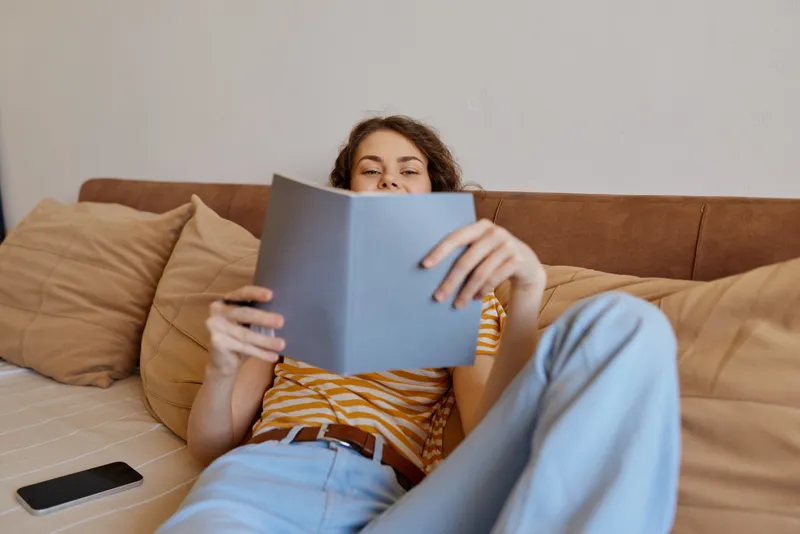 Young woman relaxing on a cozy couch reads a book in a sunlit living room, smiling softly as she leans back among plush cushions and warm natural tones.