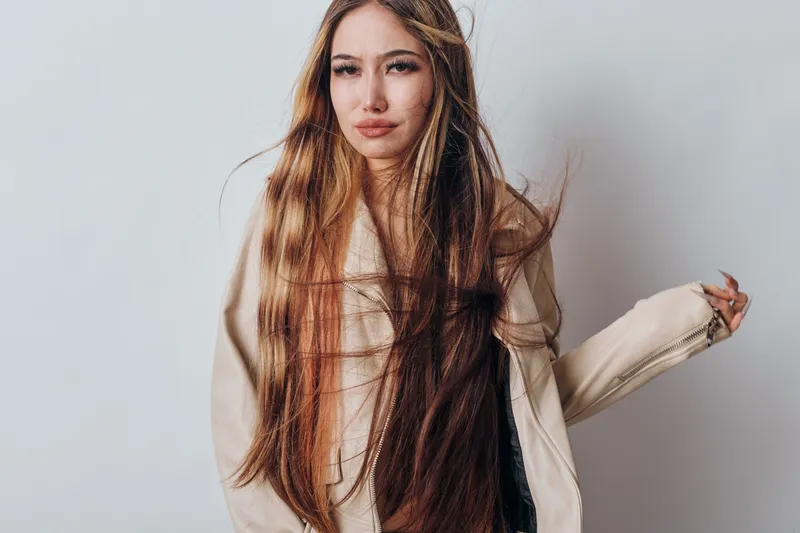 Young woman with long straight hair wearing beige jacket standing against white background, posing with calm facial expression and natural makeup in studio portrait.