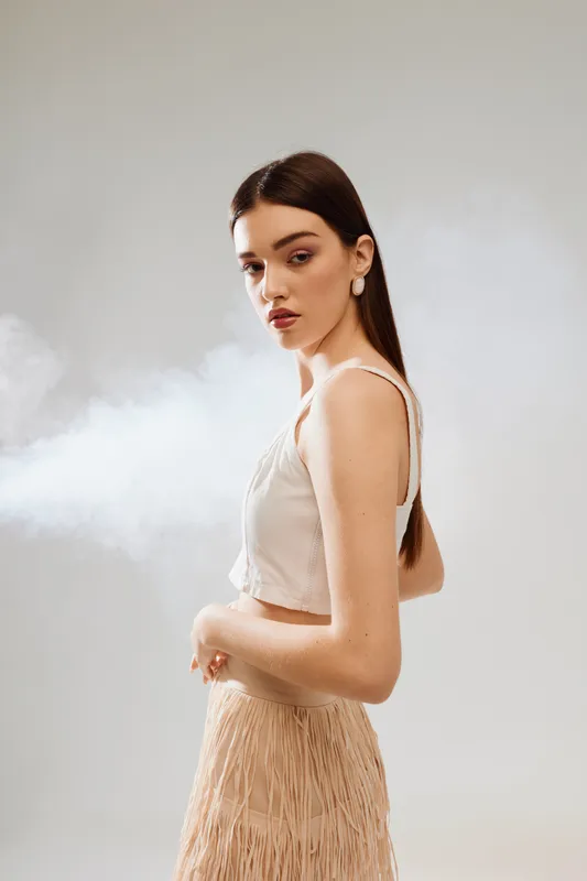 Young woman with long brown hair poses in beige sleeveless top and fringed skirt against light background with smoke. Concept of beauty, fashion, and elegance in studio setting.