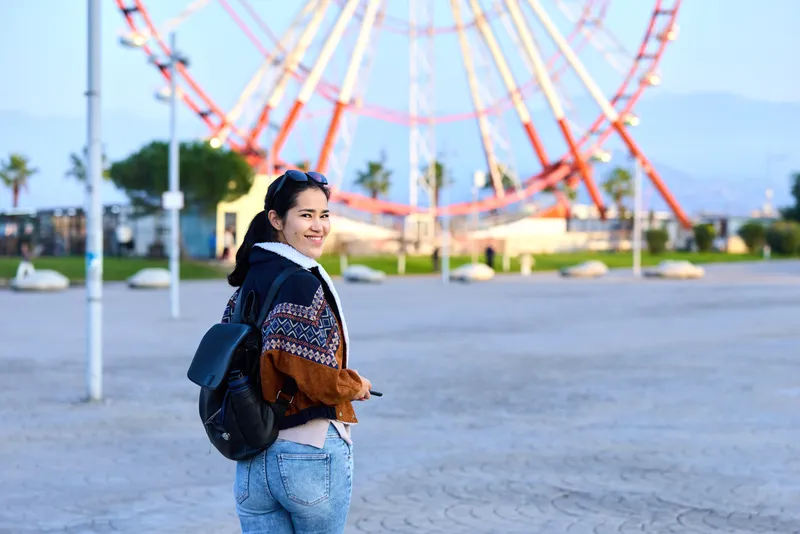 Girl backpack amusement park roller coaster outdoor casual youth smiling as she strolls near the ride, capturing a bright, airy moment of exploration and leisure in a sunny outdoor setting