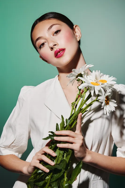 Beautiful young woman with high fashion makeup holds white daisies. She wears a white outfit against a pastel green background. Creative beauty concept focused on flowers.