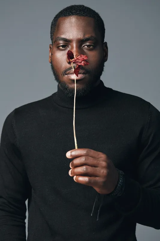 African man with dark skin wearing black turtleneck holding dried flower in front of face in creative conceptual visual style with neutral background and serious expression.