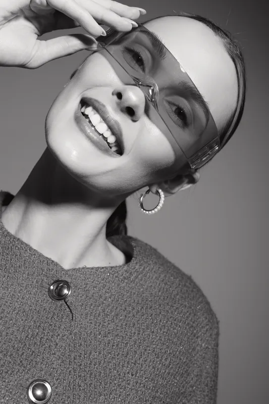 Smiling young white woman with pale skin wearing oversized sunglasses and hoop earrings in studio lighting. Fashion portrait with modern accessories and casual style.