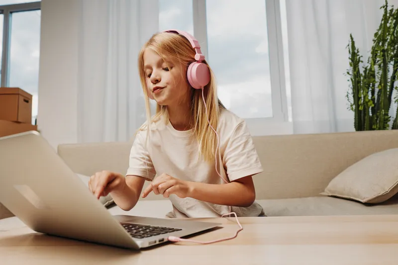 Young girl with pink headphones uses laptop on wooden table in bright living room. Casual home environment with natural light and green plant in background.